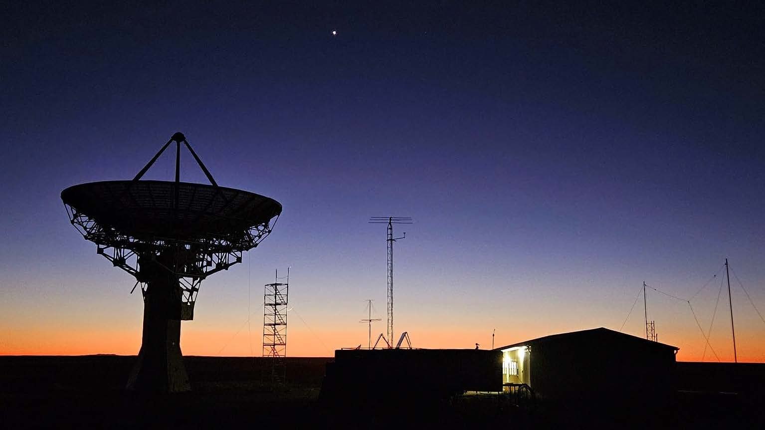 The DSES 60-foot radio telescope silhouetted against a twilight sky at the Haswell, Colorado site, with the operations building lit beside it.